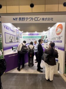 Trade show booth with purple and white panels, displays, and people conversing near marketing banners in Japanese text.